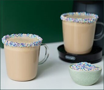 Two mugs of creamy coffee with rims decorated with colorful sprinkles and a small bowl of sprinkles